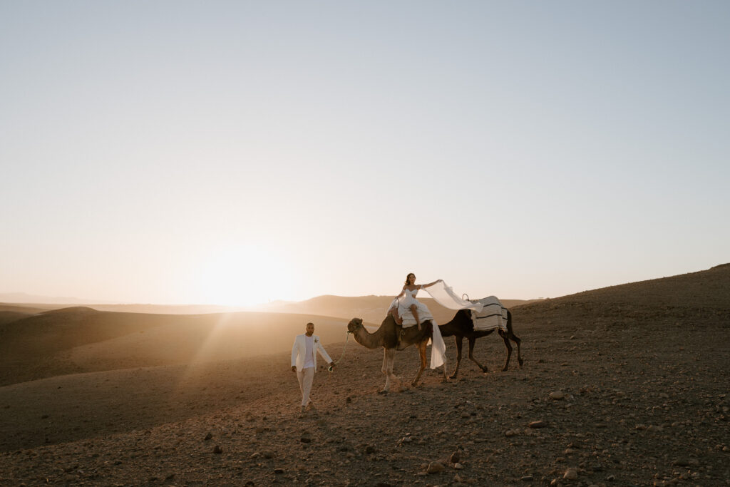 bride and groom eloping riding camel into sunset on sand dune in the agafay desert in marrakesh