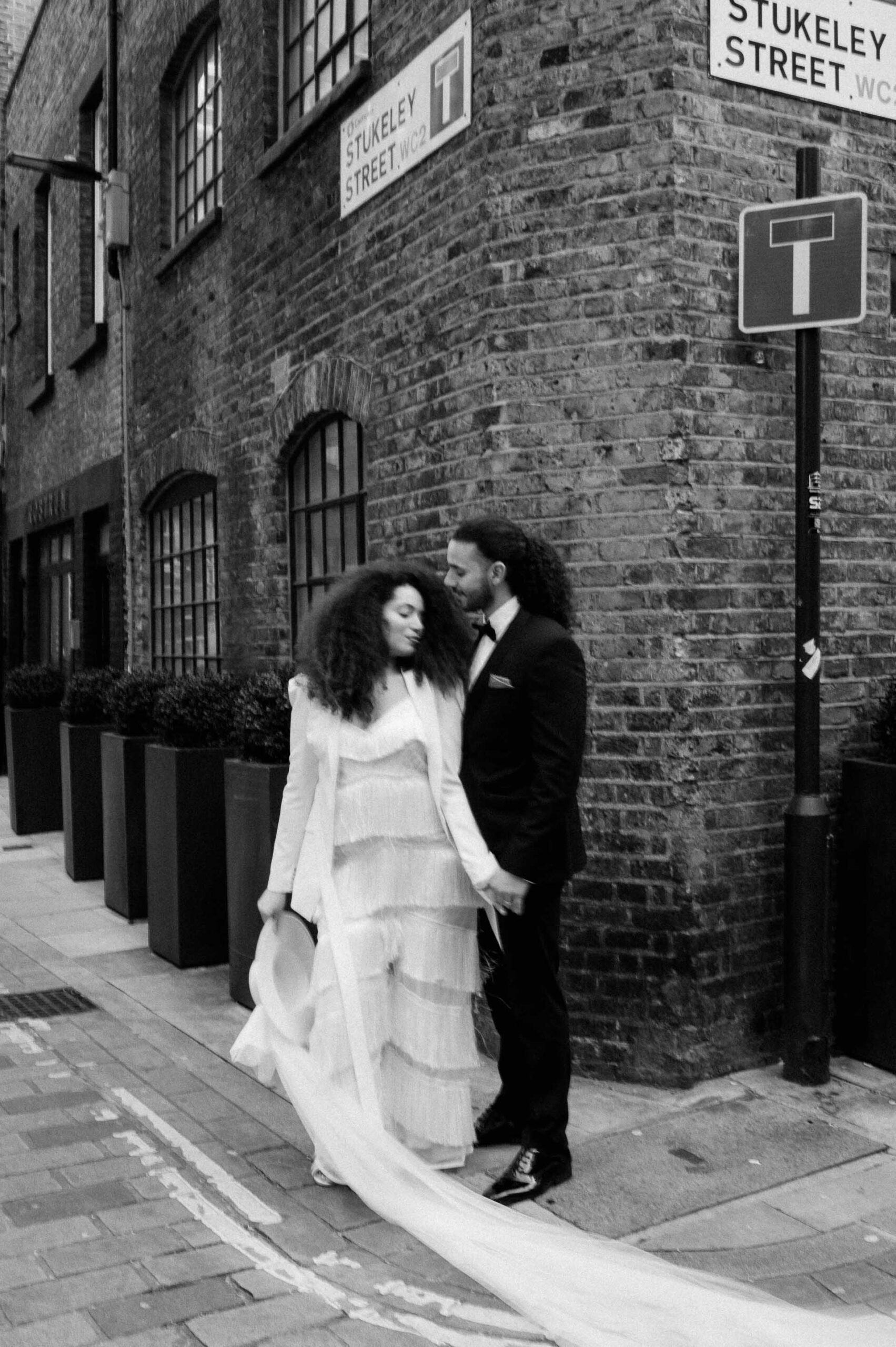 bride wearing white jumpsuit and holding white hat holding hands with groom in streets of London