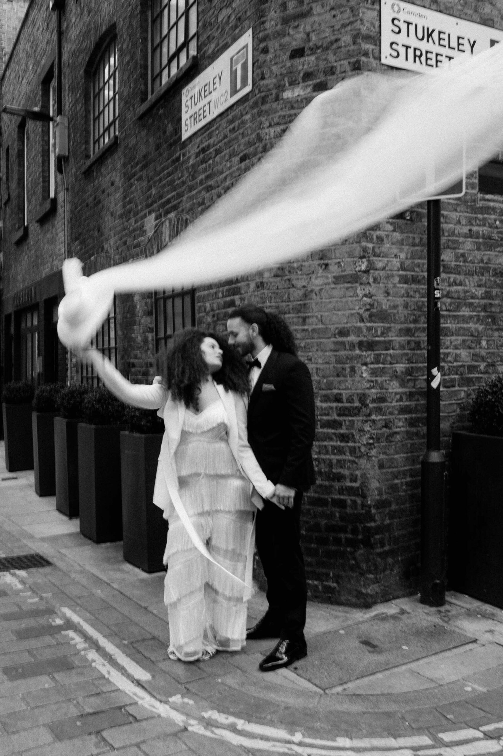 bride wearing white jumpsuit and holding white hat holding hands with groom in streets of London
