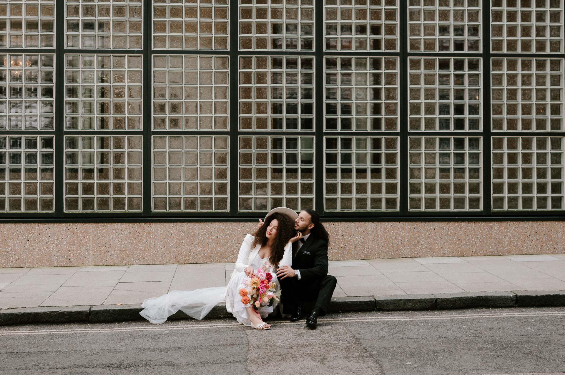 bride and groom sitting on pavement cuddling with colourful bouquet