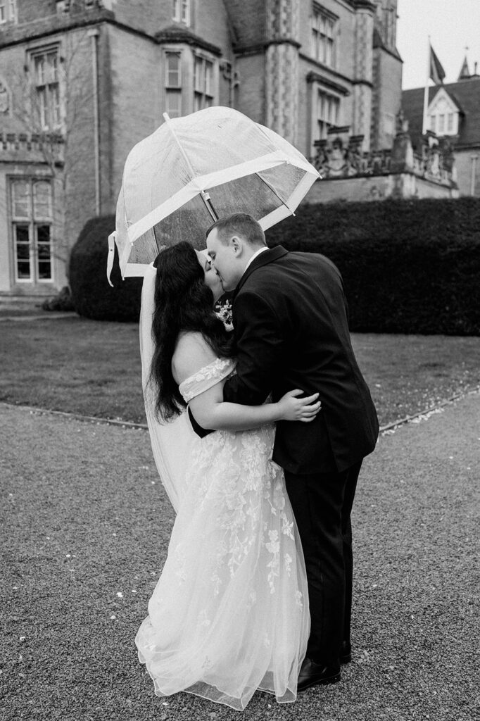 bride and groom sharing a kiss with clear umbrella walking in the rain at tortworth court